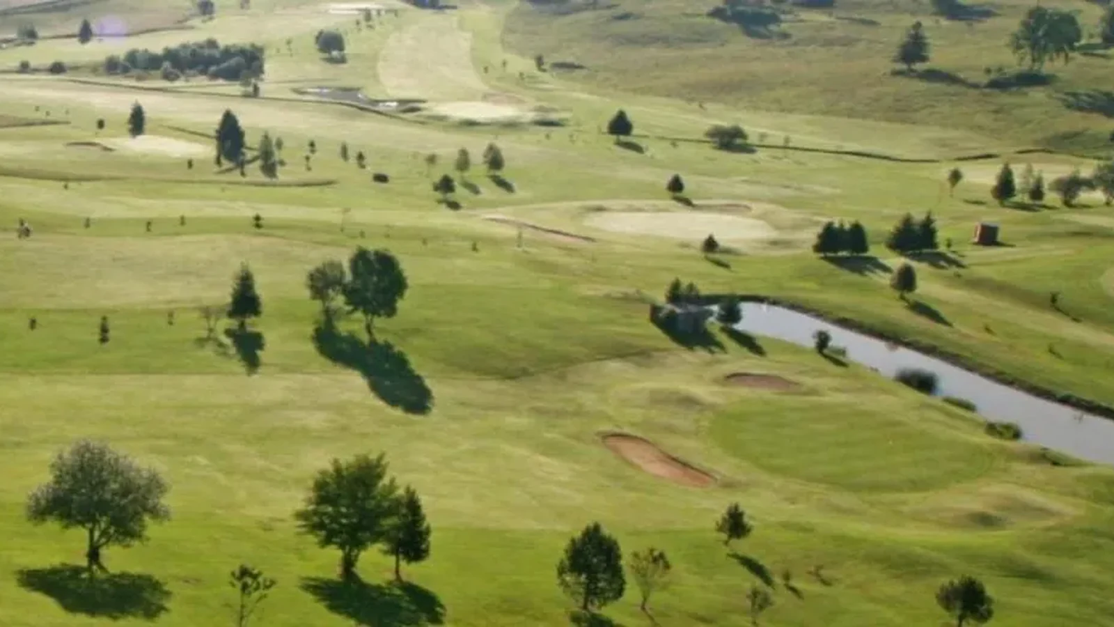 Vue aérienne du parcours vallonné, fairways, arbres et pièce d’eau ; pas de bâtiment dominant. Photographie finale inspectée sur la planche contact : pas de texte superposé ni filigrane visible ; recadrage non destructif et sujet golf représentatif. Identité exacte étayée par la page officielle, non inférée des pixels seuls. — Golf du Domaine du Mont-Saint-Jean