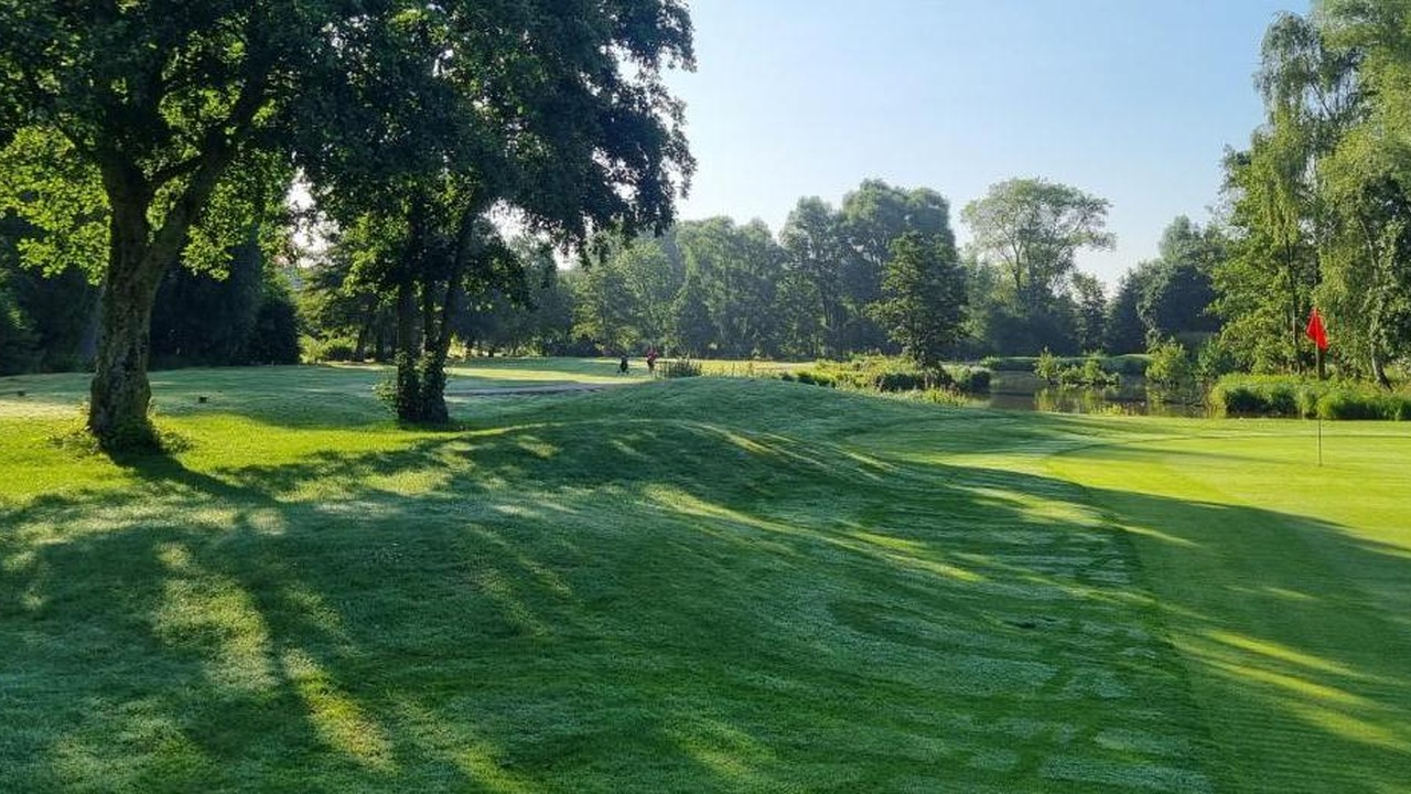 Green avec drapeau rouge et fairway ombragé par de grands arbres au Golf d’Arras.