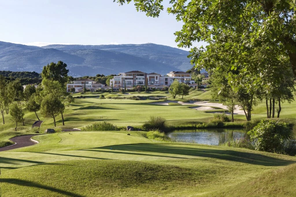 Fairways vallonnés, bunkers et pièce d’eau avec bâtiments du resort en arrière-plan.