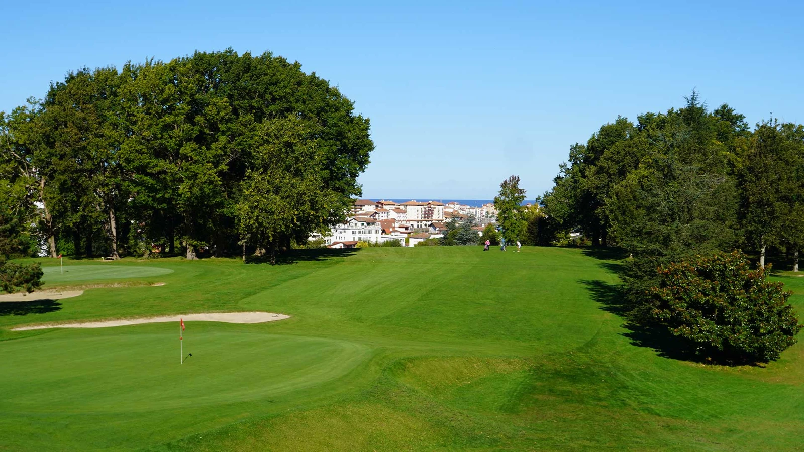 Green, bunker et fairway arboré au Golf de la Nivelle, avec les bâtiments de la côte en arrière-plan.