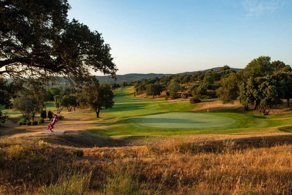 Green et fairway du Golf de Roquebrune entre arbres et collines, avec une sculpture au bord du parcours.